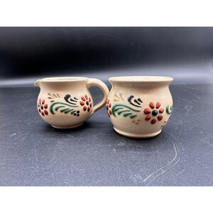 May include: A cream-colored ceramic creamer pitcher and a matching sugar bowl, both featuring hand-painted floral motifs in dark red, black, and green. The creamer has a curved handle and a small spout, while the sugar bowl has a wide opening.