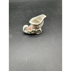 May include: A small, white ceramic shoe-shaped vase with gold trim and a pink rose accent. The vase has a curved opening and is likely vintage, with a decorative design.