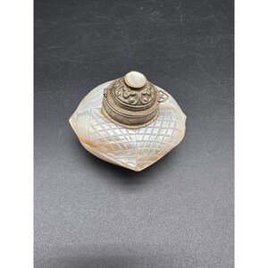 Puede incluir: Un pequeño tintero de nácar iridiscente con un patrón geométrico de diamantes en su cuerpo. El recipiente está rematado con una tapa circular de metal plateado desgastado que muestra un intrincado relieve floral y una incrustación central.