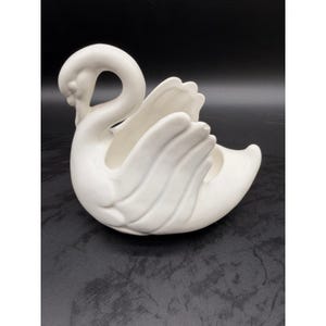 May include: A white ceramic swan-shaped planter or decorative bowl. The swan is depicted with its neck curved gracefully forward and its wings partially raised, creating a hollow centre for holding small items. The finish is smooth and matte white.