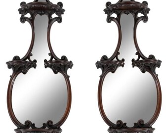 May include: A pair of antique, ornately carved wooden wall mirrors with shelves. The mirrors are oval shaped and have a dark brown finish.