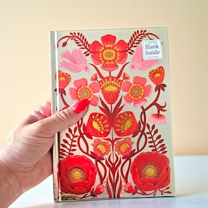 May include: A hardcover notebook with a floral design. The cover features red flowers, pink birds, and green leaves on a light beige background. The notebook has a sticker that says "Blank Inside".