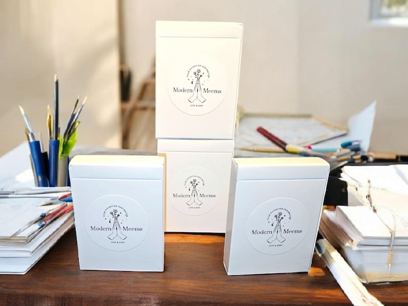 May include: Three white rectangular boxes stacked and arranged on a surface. Each box features a circular label with a black line drawing of hands holding flowers and the text Modern Meems, Est 2022, printed in a clean, black sans-serif font.