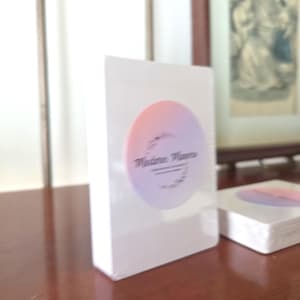 May include: A white rectangular deck of cards encased in clear plastic wrap. A circular sticker is centered on the front, featuring a gradient from soft orange to light purple. The text "Madina Muna" is printed in black script within the circle.