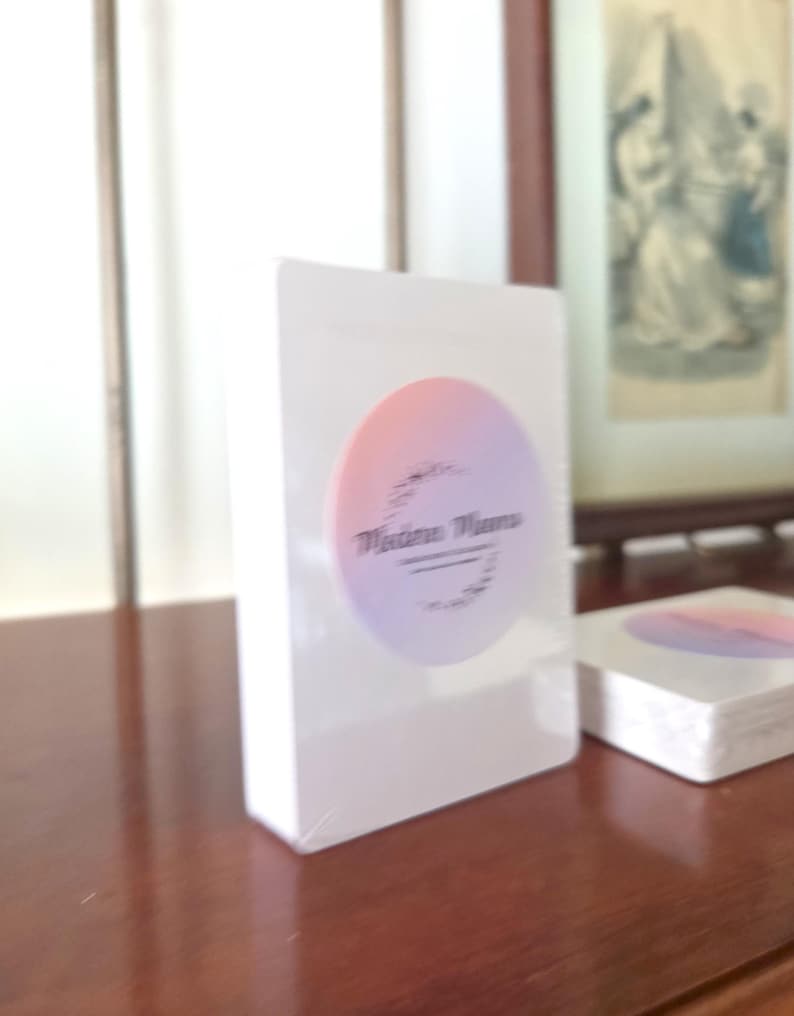 May include: A white rectangular deck of cards encased in clear plastic wrap. A circular sticker is centered on the front, featuring a gradient from soft orange to light purple. The text "Madina Muna" is printed in black script within the circle.