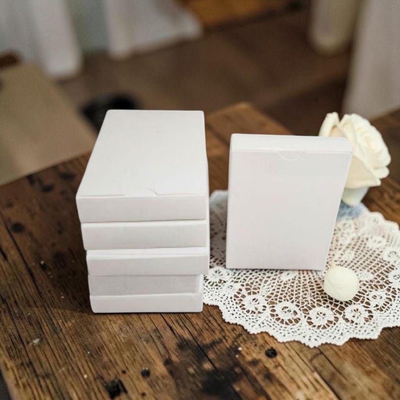 May include: A stack of four rectangular white boxes next to a single upright white box. The boxes are plain with a smooth matte finish and a small semi-circular tab on the top edge. A white lace doily and a white rose are visible nearby.