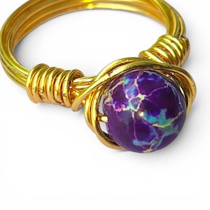 May include: A gold-toned wire-wrapped ring featuring a round, purple gemstone with blue and white veining. The ring's band and setting are crafted from coiled wire, creating a textured and artistic design. The gemstone is the focal point.