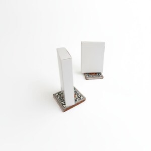May include: Two white rectangular upright blocks, each mounted on a square base featuring a decorative black and white scrollwork pattern with a central orange accent. The objects are positioned at an angle against a plain white background.