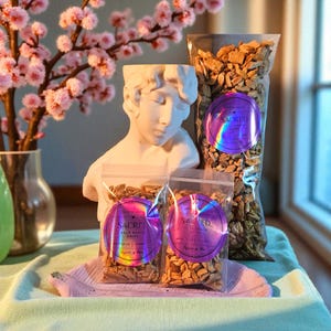 Puede incluir: Tres bolsas de plástico transparente llenas de virutas de madera marrón claro, cada una con una etiqueta holográfica circular con el texto SACRED PALO SANTO CHIPS. Las bolsas están dispuestas frente a un busto blanco sobre una bandeja morada.