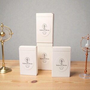 May include: Three rectangular white boxes stacked and arranged, each featuring a circular label with a black line drawing of hands holding flowers and the text Modern Merms. The boxes are positioned between two metallic decorative stands.