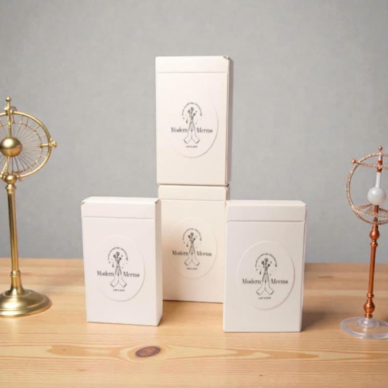 May include: Three rectangular white boxes stacked and arranged, each featuring a circular label with a black line drawing of hands holding flowers and the text Modern Merms. The boxes are positioned between two metallic decorative stands.