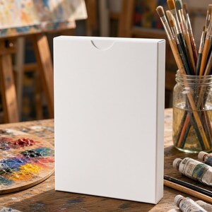 May include: A rectangular white box with a thumb notch cutout at the top center. The box is standing upright on a wooden surface surrounded by various art supplies including paintbrushes in a glass jar, paint tubes, and a wooden palette.