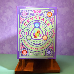 May include: A boxed crystal kit with a green and purple design. The box reads "CRYSTALS" in pink lettering, with the text "ILLUSTRATED GUIDE AND 7-PIECE CRYSTAL KIT" below. The box is displayed on a wooden stand against a purple background.