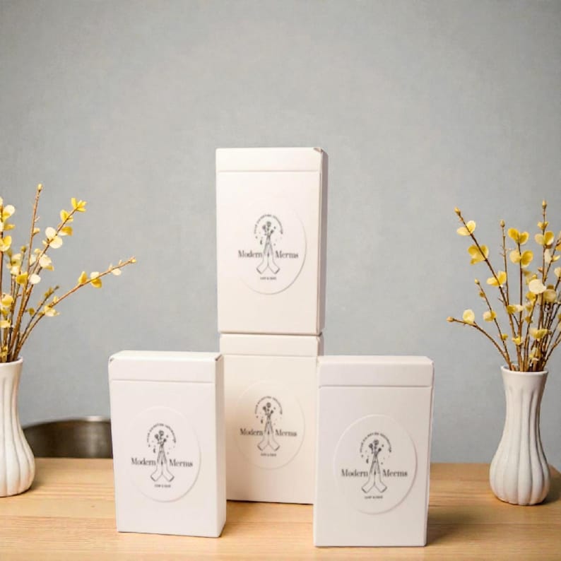May include: Three white rectangular boxes stacked in a pyramid formation, each featuring a circular label with a line drawing of hands holding flowers and the text Modern Mernas. The boxes are arranged on a wooden surface between two white vases.