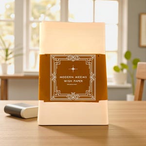 Può includere: Un pacco rettangolare di carta dei desideri Modern Meems. La confezione è per lo più bianca con una fascia marrone con testo bianco ed elementi decorativi. Un piccolo oggetto grigio e bianco si trova a sinistra della confezione su una superficie di legno.