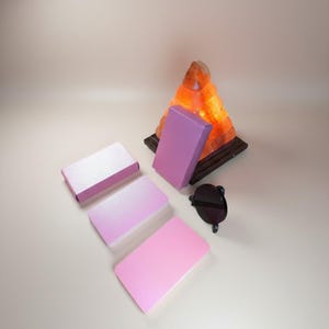 Könnte beinhalten: Eine Anordnung von rechteckigen Schachteln in Rosa- und Lilatönen, mit einer pyramidenförmigen Himalaya-Salzlampe im Hintergrund. Eine schwarze Sonnenbrille liegt daneben. Die Schachteln variieren von hellrosa bis zu einem tieferen Lila.