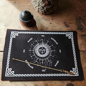 May include: A black rectangular divination mat featuring a white printed sun and moon design with the words YES, NO, MAYBE, and REPHRASE. A gold-toned metal pendulum on a delicate chain rests diagonally across the mat, which has a decorative border.