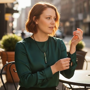 May include: A woman wearing a dark green blouse with a black necklace featuring a green and gold pendant. The blouse has long sleeves with buttoned cuffs. The background shows an outdoor cafe setting.