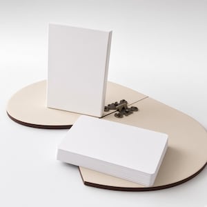 May include: A stack of blank white rectangular cards and a single upright white card positioned on a light beige, heart-shaped wooden base. The base features a dark brown edge and is connected by a decorative, ornate bronze-colored metal hinge.