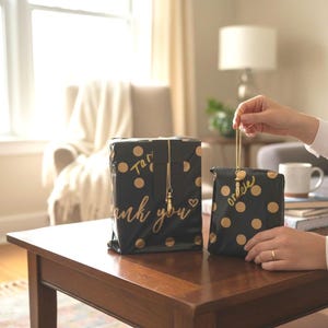 Op de afbeelding: Twee zwarte geschenkdozen met gouden stippen en gouden tekst met de tekst "thank you" en "oracle". De dozen zijn vastgebonden met gouden draad en bedels. De dozen staan op een donkerbruine houten tafel.