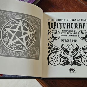 May include: An open book titled "The Book of Practical Witchcraft" by Pamela Ball. The left page has a grey pentagram design. The right page features the title in black with decorative illustrations.