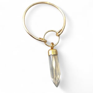 Puede incluir: Un anillo dorado con un pequeño colgante adjunto. El colgante tiene una forma clara, similar a un cristal, con una tapa dorada. El anillo es circular y tiene un diseño simple y elegante. El colgante mide aproximadamente 3,8 cm de largo.