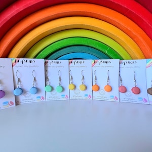 May include: Eight pairs of polymer clay earrings shaped like macarons in various colours, including light blue, green, yellow, orange, pink, and brown. Each pair is displayed on a white card with the text "TinySnaks" and "polymer clay jewellery".