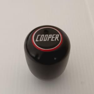 May include: Black gear shift knob with a red and white circle around the word "COOPER" in white lettering.