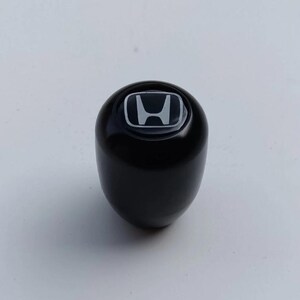Puede incluir: Un pomo de palanca de cambios esférico, negro y liso, que presenta un emblema plateado con el logotipo de Honda centrado en la parte superior. El objeto tiene un acabado brillante y una construcción sólida y redondeada.