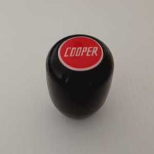 Puede incluir: Pomo de cambio de marchas negro con un círculo rojo y la inscripción "COOPER" en blanco.