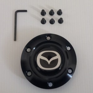May include: Black car part featuring a silver Mazda logo, accompanied by six black screws and a black Allen key. The part has six holes around the edge.