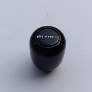 May include: A black gear stick knob with the word "nismo" in white lettering and a red "o" on the top. The gear stick knob is a rounded, cylindrical shape and is set against a white background.