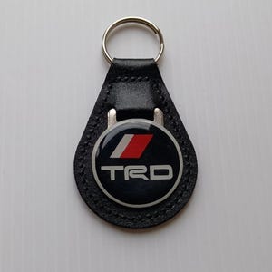 Puede incluir: Llavero de cuero negro con un llavero plateado. El llavero presenta un emblema circular con las letras "TRD" en blanco, una franja roja y blanca y un fondo negro.