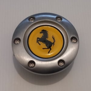 Könnte beinhalten: Ein silberfarbenes, kreisförmiges Emblem mit einem gelben Zentrum, das ein schwarzes, aufbäumendes Pferd-Logo zeigt. Das Emblem ist mit sechs kleinen Schrauben befestigt. Autozubehör.
