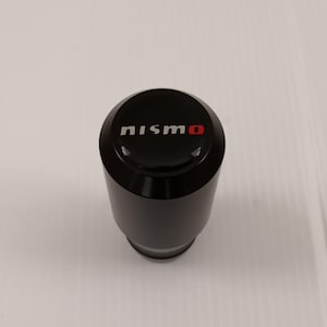 以下が含まれることがあります： 白と赤の文字で「nismo」という文字が入った黒いシフトノブ。 シフトノブは円筒形で、上部が丸く、光沢のある仕上がりです。 この製品はおそらく自動車部品です。