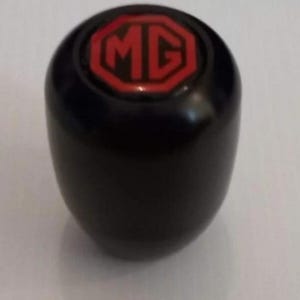 Puede incluir: Un pomo de palanca de cambios cilíndrico de color negro con una superficie superior lisa y redondeada. En el centro hay un emblema octogonal con fondo rojo y las letras negras MG impresas en negrita, rodeado por un borde de color negro.