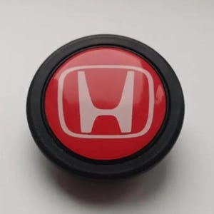 Pu&ograve; includere: Emblema Honda rotondo, rosso con logo 'H' bianco, racchiuso in un anello nero. Probabilmente un componente o accessorio per auto.