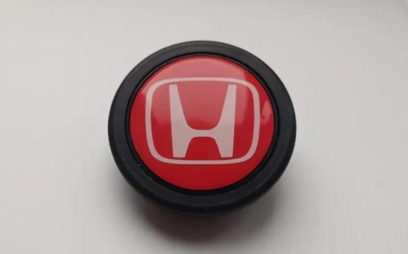 Pu&ograve; includere: Emblema Honda rotondo, rosso con logo 'H' bianco, racchiuso in un anello nero. Probabilmente un componente o accessorio per auto.