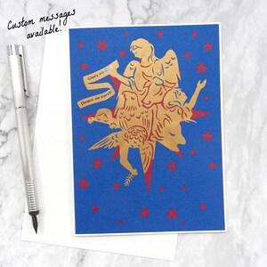 Op de afbeelding: Een blauwe kaart met een gouden silhouet van een engel met een duif en een ster. De engel houdt een rol papier vast met de tekst "Eer aan God" en "Vrede op aarde".