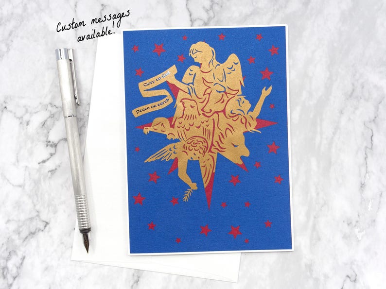 Op de afbeelding: Een blauwe kaart met een gouden silhouet van een engel met een duif en een ster. De engel houdt een rol papier vast met de tekst "Eer aan God" en "Vrede op aarde".