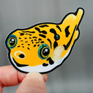 Op de afbeelding: Een gestanste sticker met een cartoon kogelvis. De vis heeft een gele body met zwarte vlekken, grote blauwe en groene ogen en een witte buik. De sticker wordt door een hand tegen een grijze achtergrond gehouden.