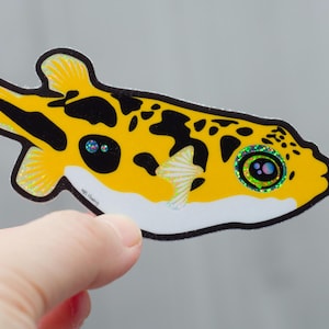 Op de afbeelding: Een gele kogelvissticker met zwarte vlekken en een witte buik. De vis heeft iriserende ogen en vinnen en wordt door een hand vastgehouden. De sticker heeft een zwarte omtrek en is waarschijnlijk voor decoratieve doeleinden.