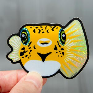 Op de afbeelding: Een gele kogelvissticker met zwarte stippen en een witte buik. De sticker heeft iriserende accenten op de vinnen en rond de ogen. De ogen zijn blauw en groen. De sticker wordt door een hand vastgehouden.