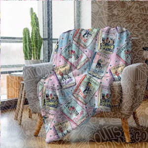 May include: A soft, patterned throw blanket draped over a chair. The blanket features a repeating design of book covers with the text "ELLE KENNEDY" and book titles like "The Deal" and "The Score" on a pastel background with stars.