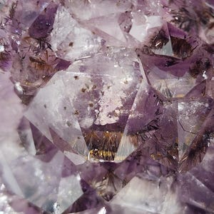 May include: Close-up of a natural amethyst crystal formation. The image showcases a complex arrangement of translucent, faceted surfaces in shades of purple and white. Fine, dark inclusions and golden accents add detail to the crystalline structure.