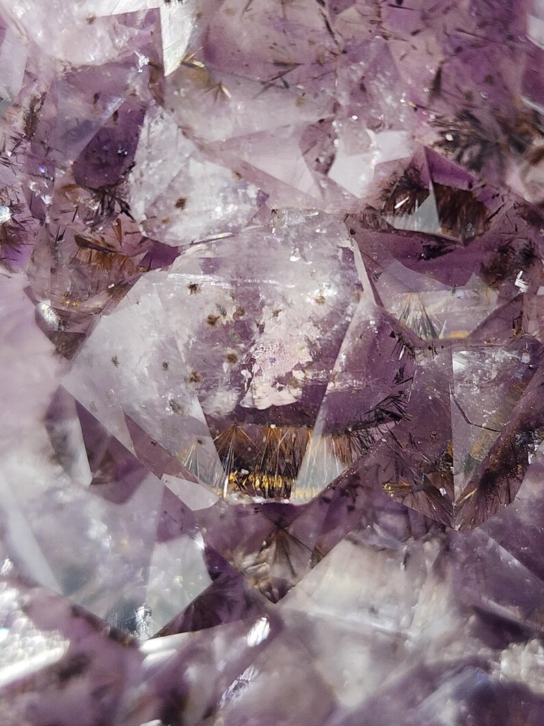 May include: Close-up of a natural amethyst crystal formation. The image showcases a complex arrangement of translucent, faceted surfaces in shades of purple and white. Fine, dark inclusions and golden accents add detail to the crystalline structure.