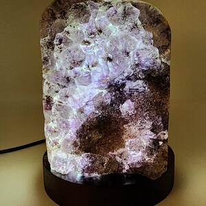 May include: A unique amethyst crystal lamp with a dark wooden base. The crystal is a mix of purple and white hues, illuminated from within. The lamp's rough, natural texture adds to its aesthetic appeal. A black power cord extends from the base.