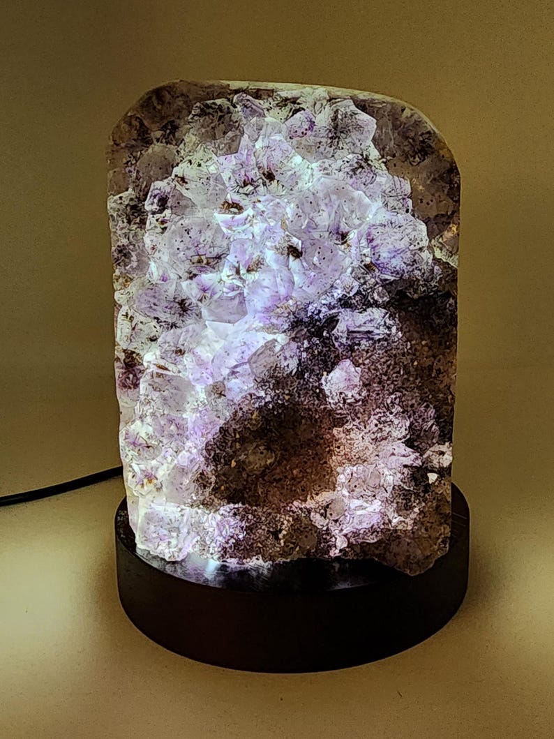 May include: A unique amethyst crystal lamp with a dark wooden base. The crystal is a mix of purple and white hues, illuminated from within. The lamp's rough, natural texture adds to its aesthetic appeal. A black power cord extends from the base.