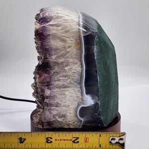 May include: A natural amethyst geode crystal lamp with a dark wood base. The crystal features shades of purple, white, and green. The lamp is approximately 4 inches tall, measured with a Stanley tape measure.