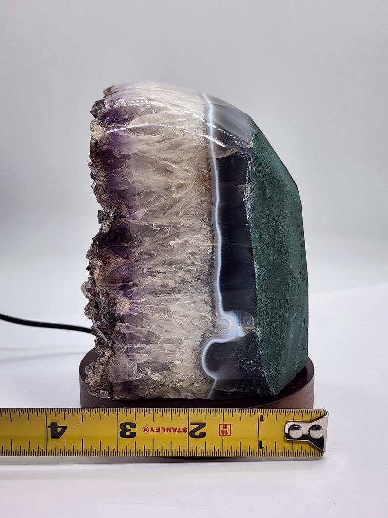 May include: A natural amethyst geode crystal lamp with a dark wood base. The crystal features shades of purple, white, and green. The lamp is approximately 4 inches tall, measured with a Stanley tape measure.